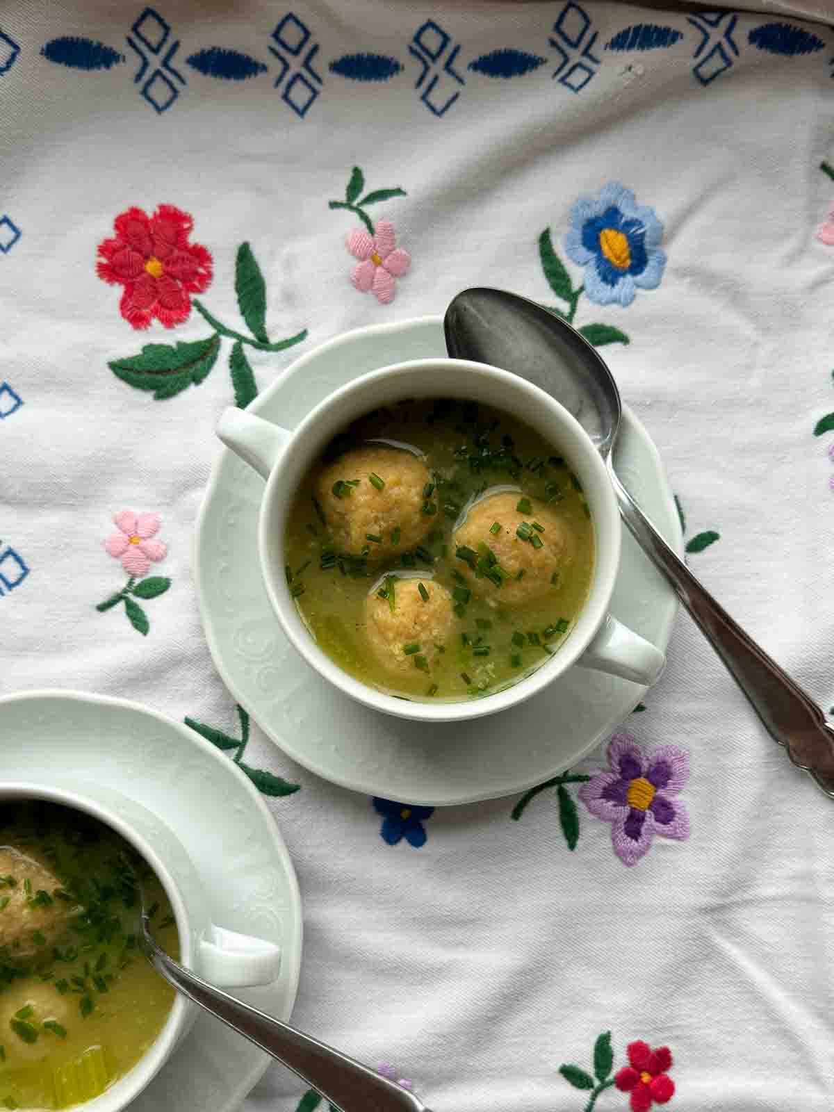 Breadcrumbs dumplings in soup cups with creamy vegetable soup on floral tablecloth.